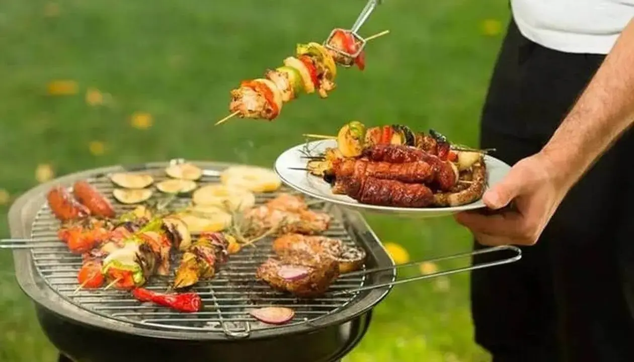 Un homme prépare un barbecue avec des brochettes et des saucisses. Qui a inventé le barbecue ? Mystère !