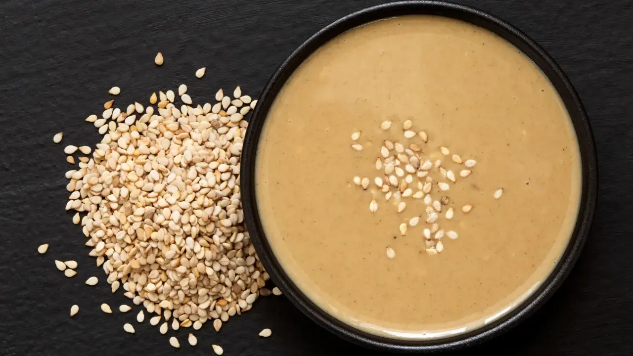 Un bol de sauce tahini onctueuse, garni de graines de sésame, à côté d'un tas de graines de sésame sur fond noir.