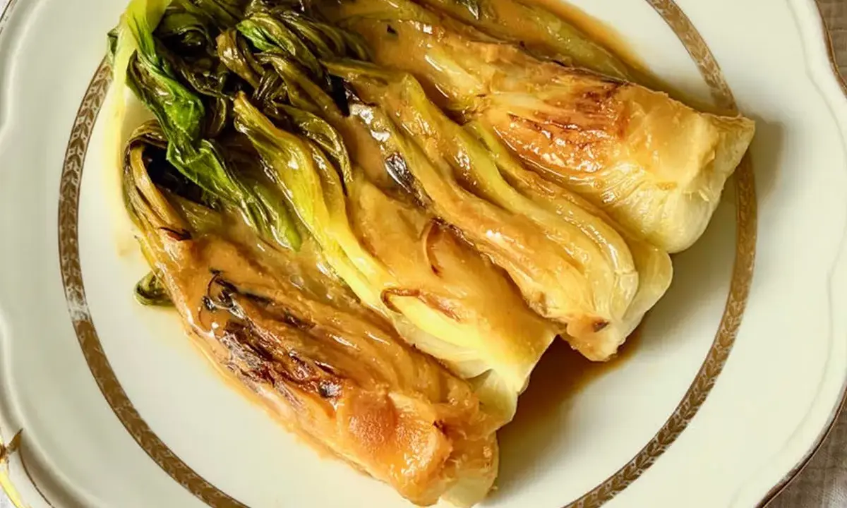 Pak choi braisé, tendre et savoureux, nappé d'une sauce dorée, servi sur une assiette élégante.