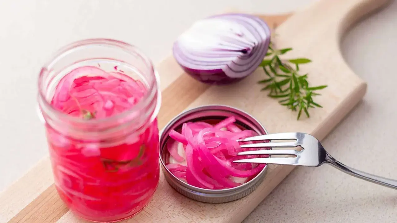 Pickles oignon rouge vibrants dans un bocal, avec une moitié d'oignon rouge et une branche de romarin sur une planche à découper.