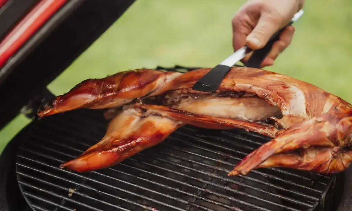 Un lapin entier est cuit sur un barbecue, sa peau dorée et croustillante. Une spatule retourne délicatement la viande pour une cuisson parfaite.