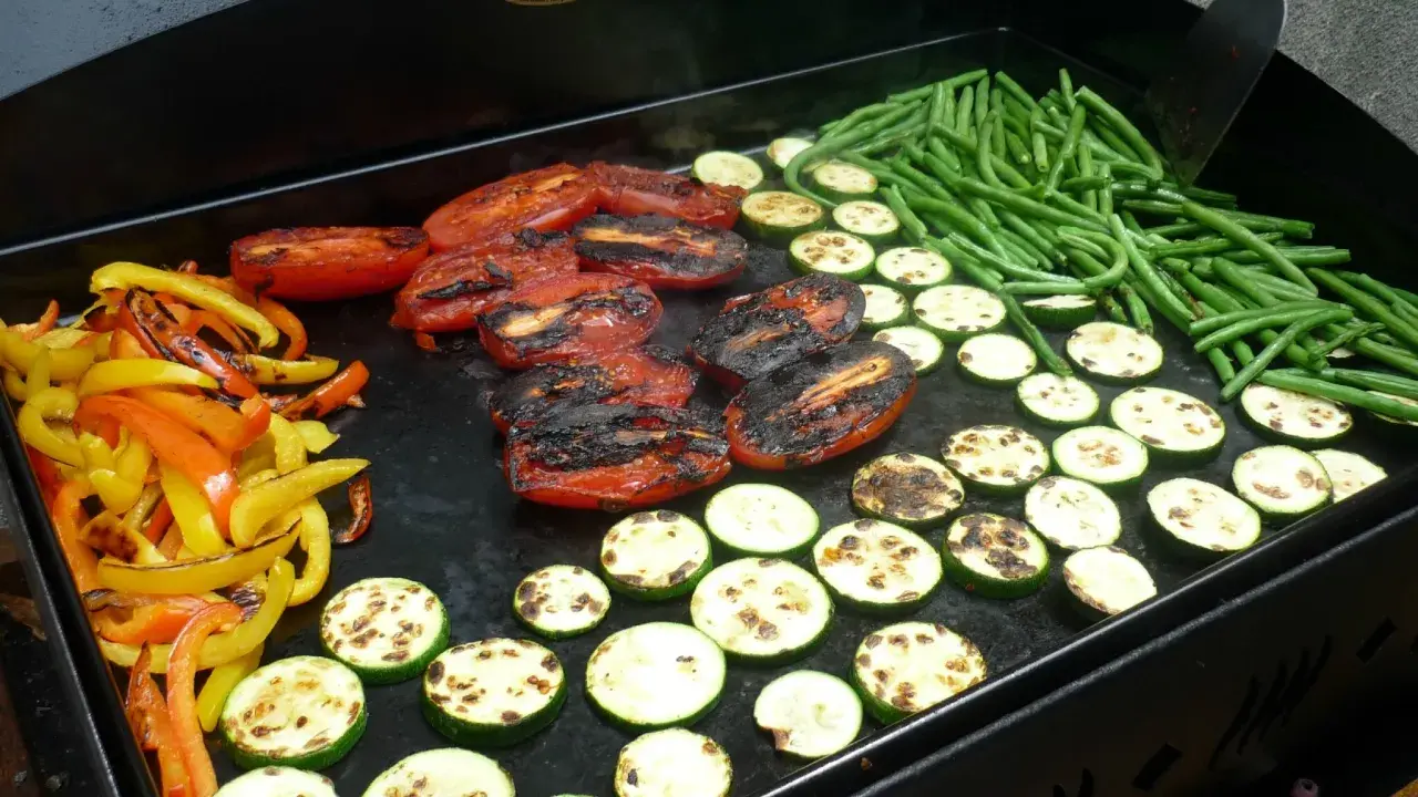 Légumes variés (poivrons, courgettes, tomates, haricots verts) en cours de cuisson sur une plancha. Le temps de cuisson des légumes à la plancha est idéal pour un repas sain et savoureux.