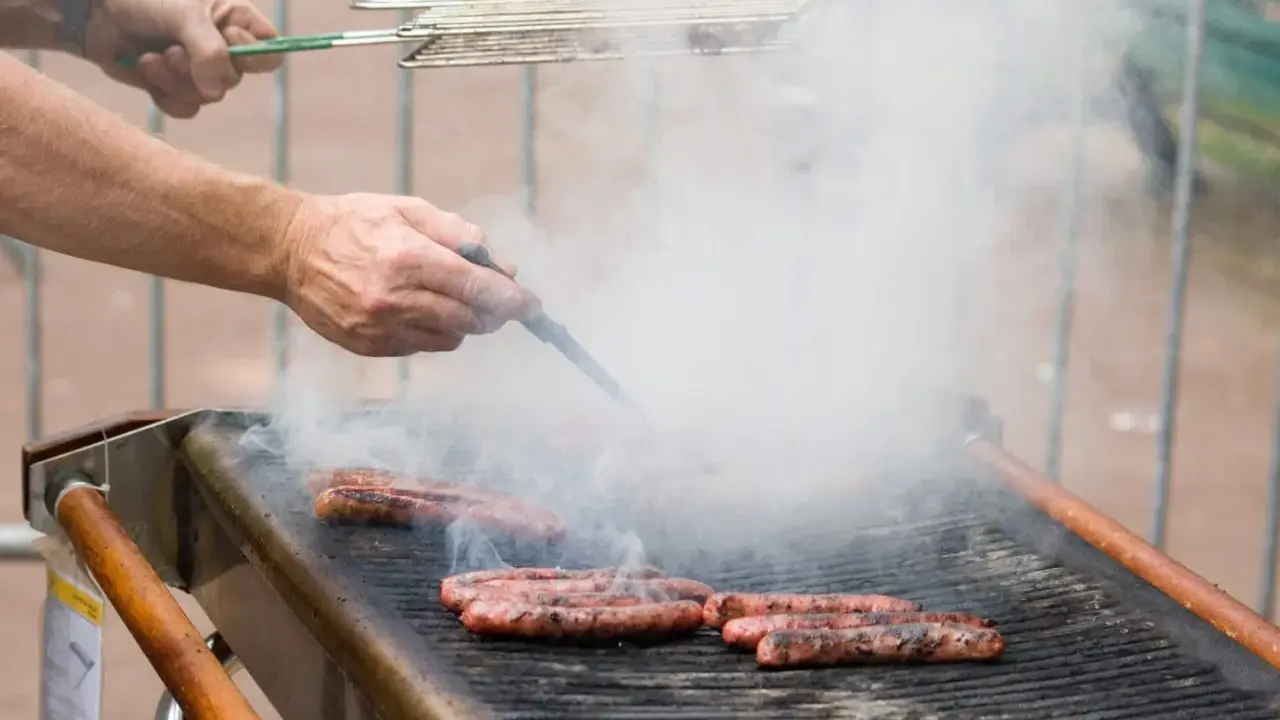 Des merguez grillent sur un barbecue, dégageant une fumée épaisse. Un homme les retourne avec une pince, se demandant s'il faut piquer les merguez pour une cuisson parfaite.