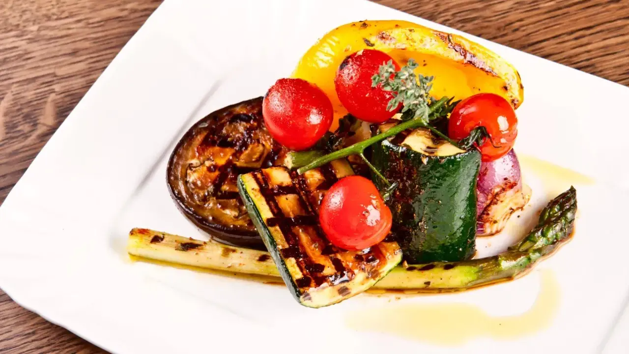 Légumes grillés : courgette, tomate, poivron jaune, aubergine, asperge et oignon rouge, arrosés d'une huile dorée.