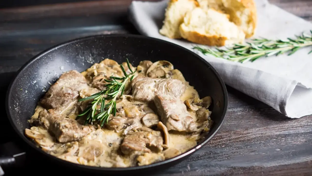 Délicieux sauté de porc crémeux aux champignons, garni de romarin frais, servi dans une poêle avec du pain croustillant à côté.