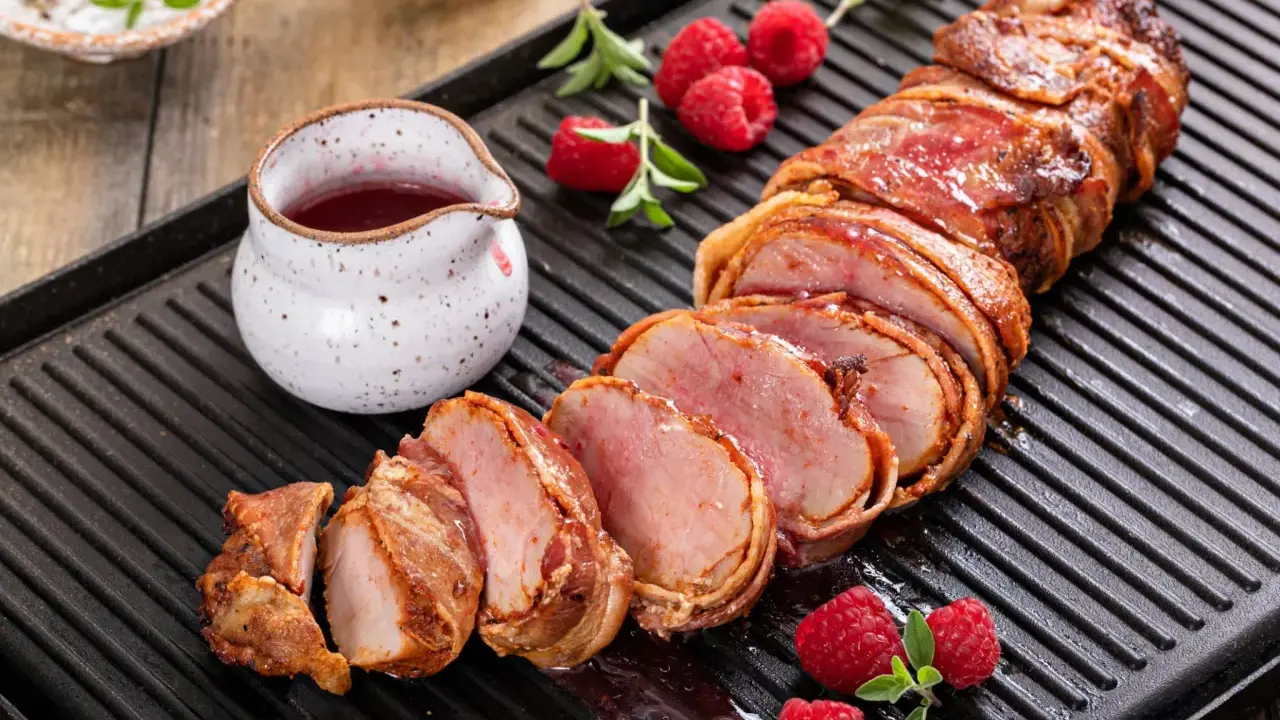Filet mignon enrobé de bacon, tranché, servi avec des framboises et une sauce rouge sur une plaque de cuisson.