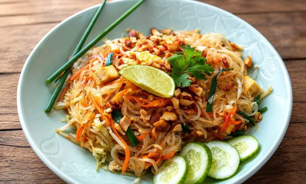 Un plat de pad thai coloré avec des nouilles de riz, des carottes, des cacahuètes concassées, du tofu, un quartier de citron vert et des rondelles de concombre.
