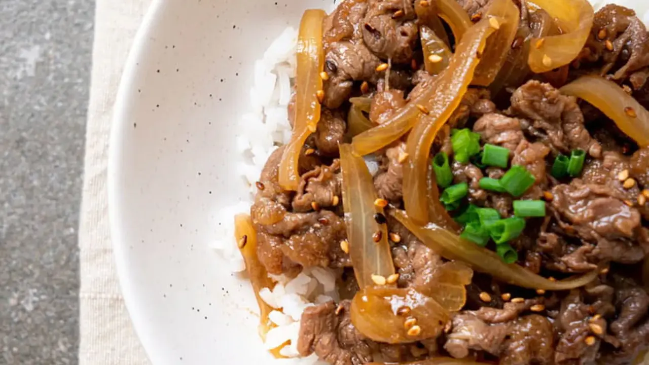 Un plat de riz blanc garni de fines tranches de bœuf aux oignons caramélisés, parsemé de graines de sésame et de ciboule ciselée. Un délice de boeuf aux oignons.