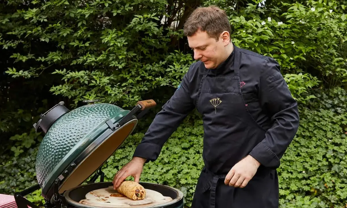 Un chef prépare un rôti sur une pierre à pizza dans un barbecue binchotan vert, entouré de verdure luxuriante.