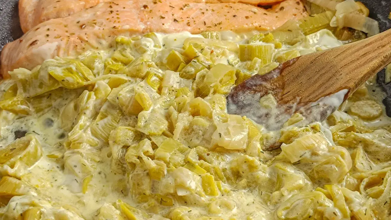 Filets de saumon poêlés sur une fondue de poireaux crémeuse, parsemée d'herbes. Une cuillère en bois remue ce plat savoureux.