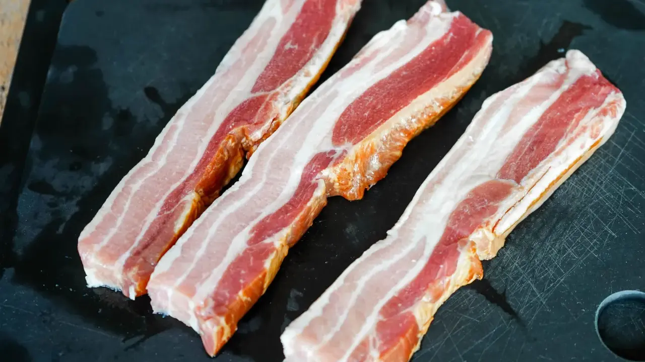 Trois tranches de lard fumé, prêtes pour une recette savoureuse, reposent sur une planche à découper noire.