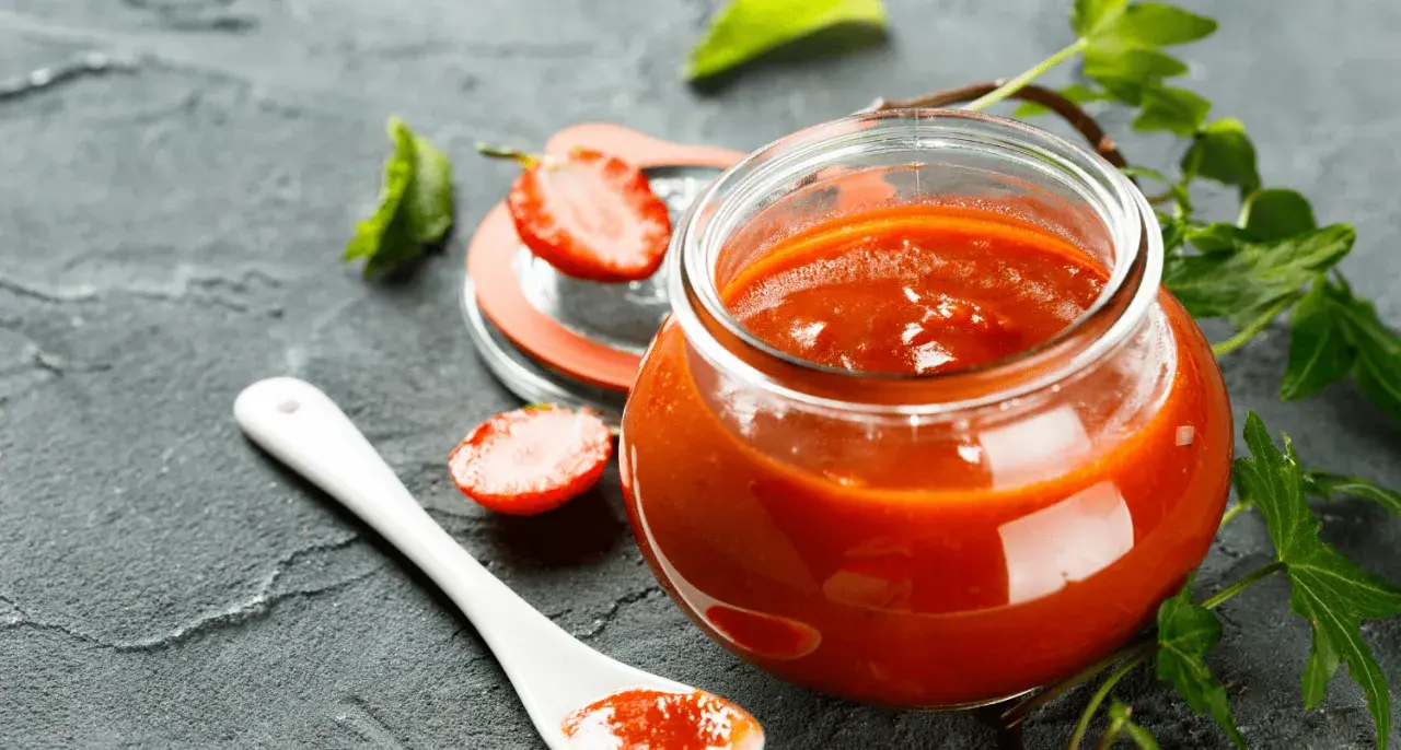 Un pot de ketchup maison, garni de fraises et d'une cuillère blanche, sur fond sombre.