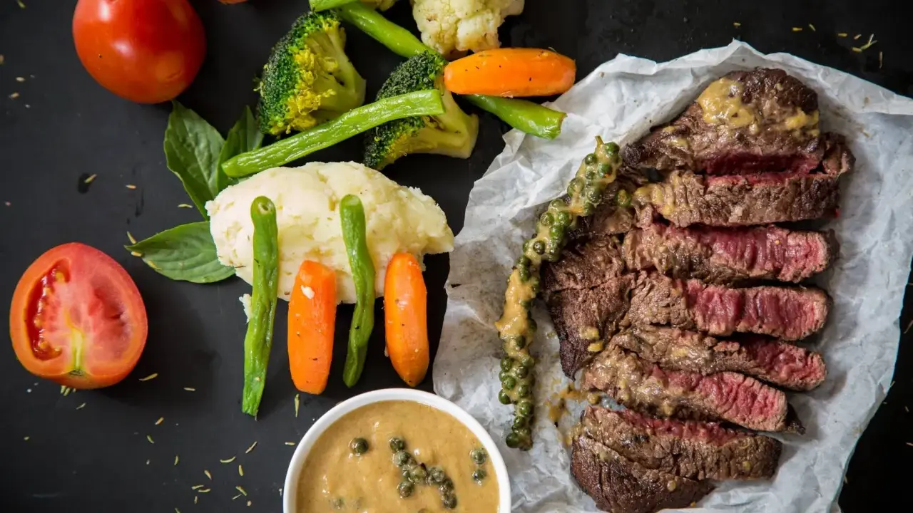 Un steak tranché, servi avec des légumes et deux types de sauces. L'une est une sauce crémeuse aux grains de poivre vert, l'autre est une sauce plus claire dans un bol.