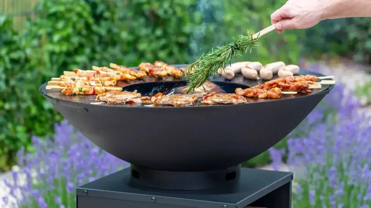Un brasero moderne cuit brochettes et saucisses. On y voit comment fonctionne un brasero, avec une branche de romarin pour parfumer.