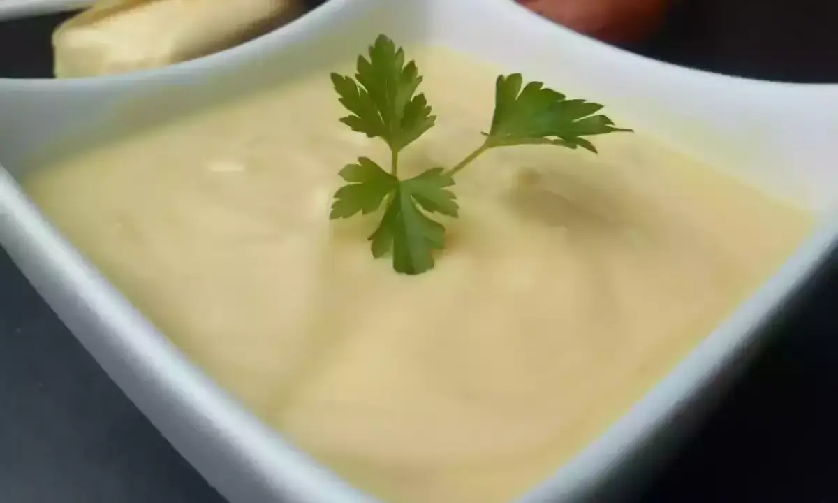 Un bol de beurre blanc crémeux, garni d'une feuille de persil. Des échalotes et un morceau de pain sont visibles en arrière-plan.
