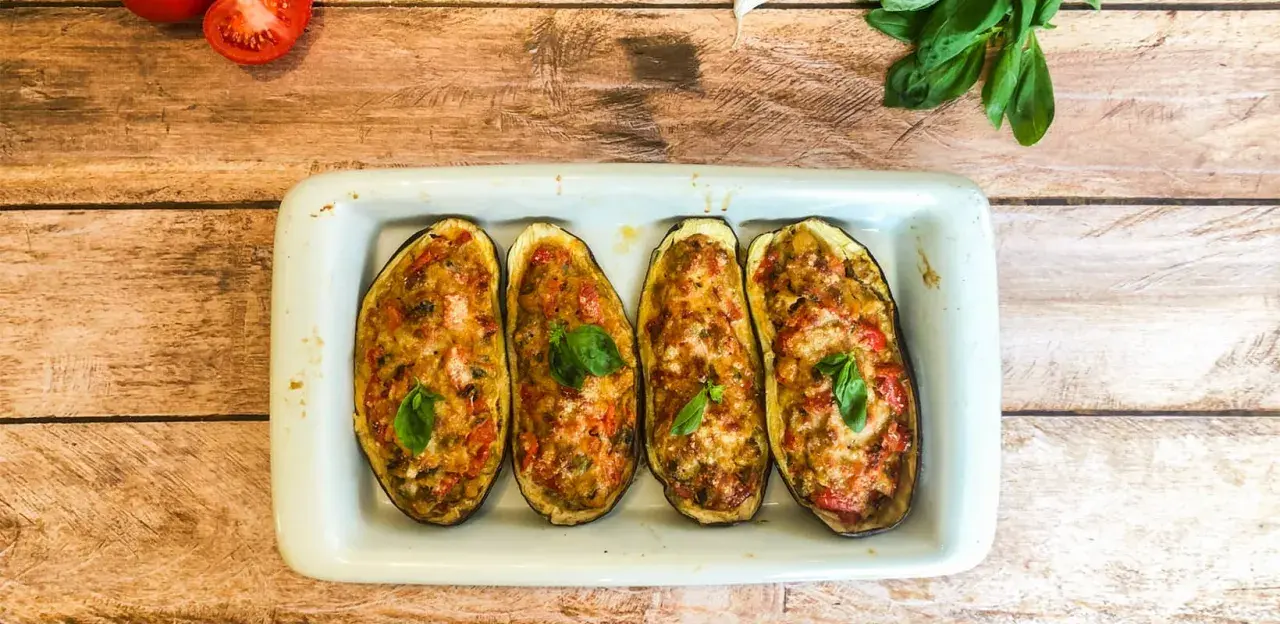 Quatre moitiés d'aubergines garnies de légumes et de fromage gratiné, décorées de feuilles de basilic frais.