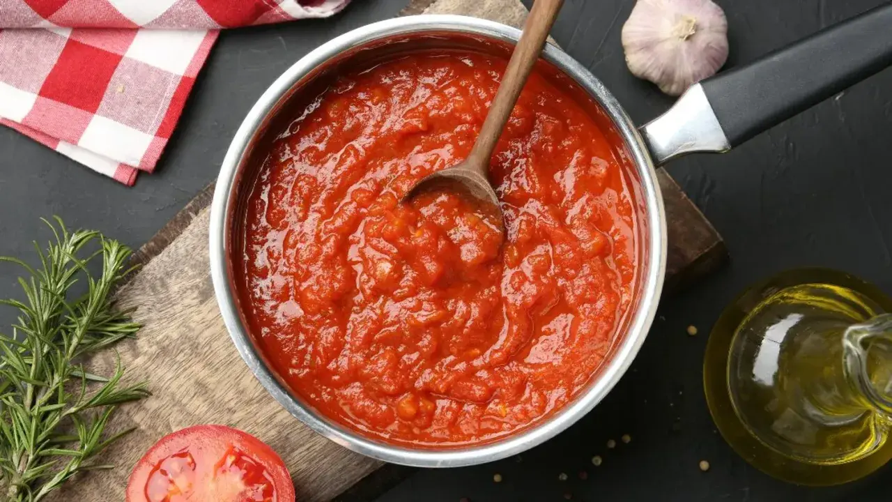 Une casserole remplie de coulis de tomates maison, avec une cuillère en bois. Des ingrédients frais comme du romarin, de l'ail et une tomate coupée en deux sont disposés autour.