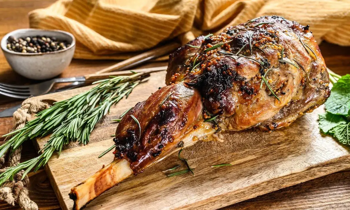 Un gigot d'agneau tendre, rôti à la perfection, est présenté sur une planche en bois avec du romarin frais et des grains de poivre.