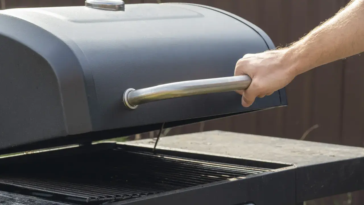 Main d'homme ouvrant le couvercle d'un barbecue noir. Apprenez comment utiliser un barbecue avec couvercle pour des grillades parfaites.
