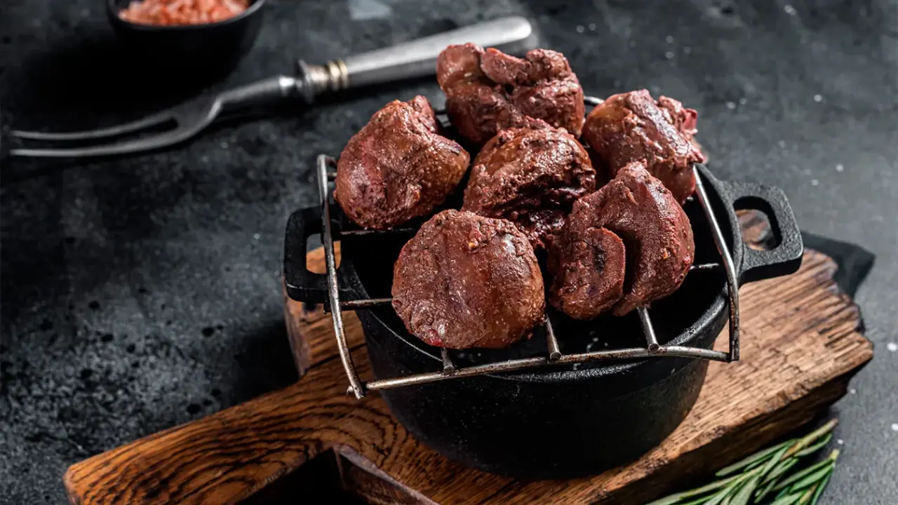 Un plat de rognons de veau grillés, servis dans une cocotte en fonte sur une planche en bois, avec une pincée de sel rose et une branche de romarin.