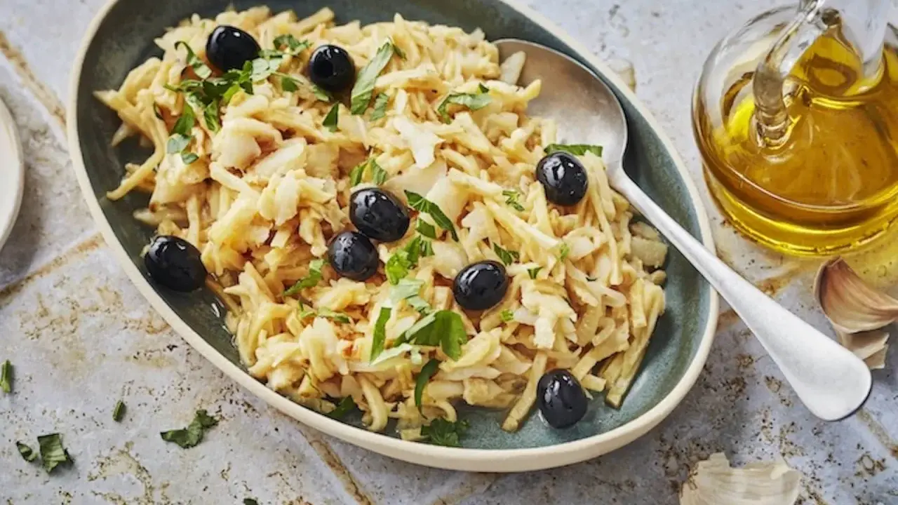 Un plat de bacalhau à brás, effiloché et garni d'olives noires et de persil frais. Une touche d'huile d'olive et d'ail est visible à côté.