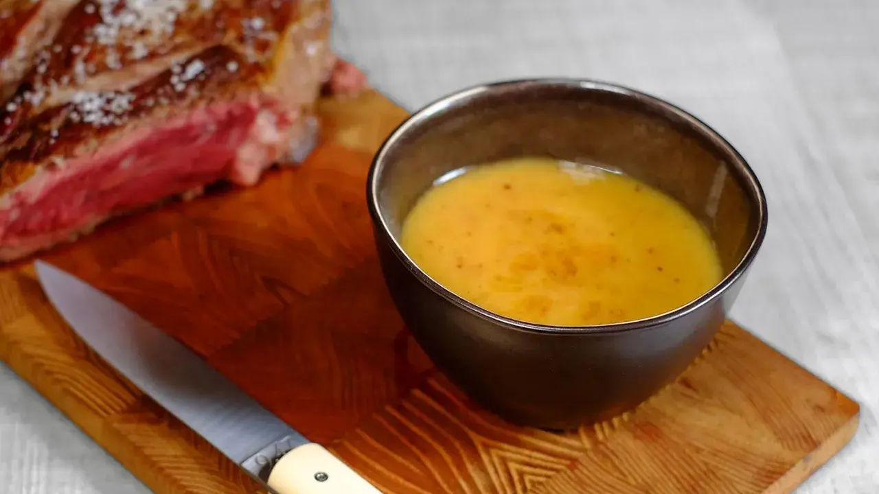 Un bol de sauce pour côte de bœuf, onctueuse et dorée, à côté d'une côte de bœuf saignante et d'un couteau sur une planche en bois.