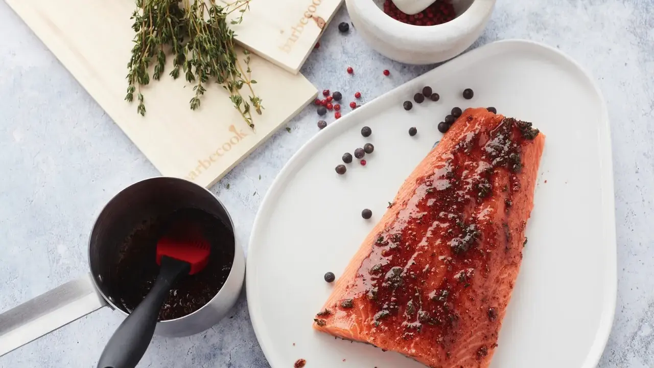 Un filet de saumon mariné est prêt à être cuit sur une planche de cèdre. Des herbes fraîches et des grains de poivre complètent la scène.