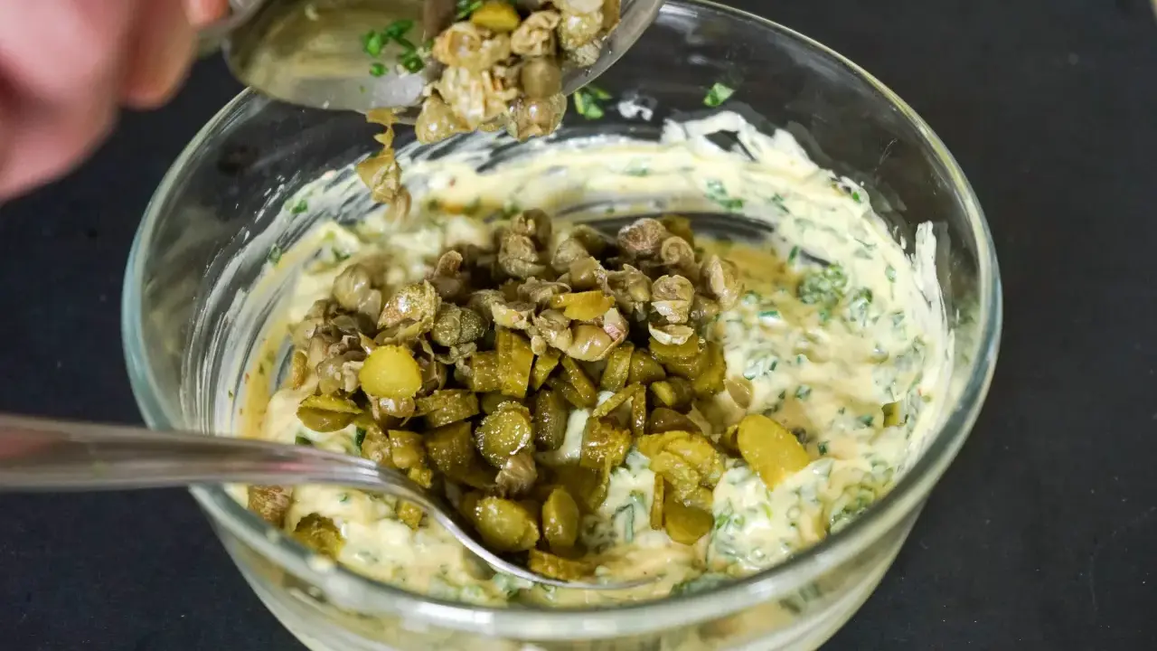 Ajout de câpres et cornichons hachés à une base crémeuse pour préparer une sauce gribiche savoureuse.