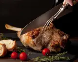 Une main utilise un couteau et une fourchette pour découper un gigot d'agneau rôti, prêt à être servi avec du pain et des tomates.