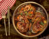 Un plat d'osso bucco mijoté, garni de persil frais, servi avec une serviette à carreaux rouges et blancs et des couverts.