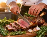 Un chef découpe un gigot d'agneau rôti, garni de romarin et d'ail. Une belle illustration pour une recette gigot agneau.
