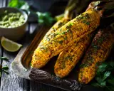 Épis de maïs grillés, parsemés d'herbes fraîches, servis sur un plateau avec une sauce verte et des quartiers de citron vert. Un délice parfait pour un repas mais au barbecue.