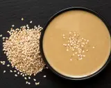 Un bol de sauce tahini onctueuse, garni de graines de sésame, à côté d'un tas de graines de sésame sur fond noir.
