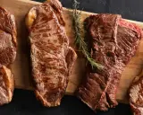 Quatre pièces de viande de saison octobre, grillées à la perfection, sur une planche en bois avec des herbes et du sel.