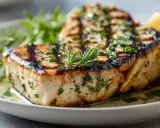 Un épais morceau d'espadon grillé, garni d'herbes fraîches et d'un quartier de citron, sur une assiette blanche.