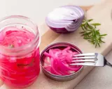 Pickles oignon rouge vibrants dans un bocal, avec une moitié d'oignon rouge et une branche de romarin sur une planche à découper.