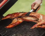 Un lapin entier est cuit sur un barbecue, sa peau dorée et croustillante. Une spatule retourne délicatement la viande pour une cuisson parfaite.