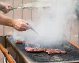 Des merguez grillent sur un barbecue, dégageant une fumée épaisse. Un homme les retourne avec une pince, se demandant s'il faut piquer les merguez pour une cuisson parfaite.