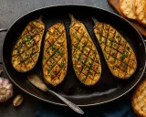 Quatre moitiés d'aubergine au four, grillées et parsemées de persil frais, dans un plat ovale avec de l'ail et des toasts grillés à côté.
