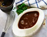 Sauce à la viande fumante dans un saucier, avec un fouet, du persil et un mortier rempli de grains de poivre.