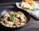Délicieux sauté de porc crémeux aux champignons, garni de romarin frais, servi dans une poêle avec du pain croustillant à côté.