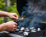 Des mains allument des charbons pour faire un barbecue. La fumée monte, annonçant un bon repas en plein air.