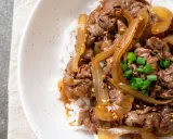Un plat de riz blanc garni de fines tranches de bœuf aux oignons caramélisés, parsemé de graines de sésame et de ciboule ciselée. Un délice de boeuf aux oignons.
