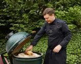 Un chef prépare un rôti sur une pierre à pizza dans un barbecue binchotan vert, entouré de verdure luxuriante.