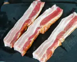 Trois tranches de lard fumé, prêtes pour une recette savoureuse, reposent sur une planche à découper noire.