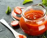Un pot de ketchup maison, garni de fraises et d'une cuillère blanche, sur fond sombre.