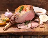 Un gigot d'agneau rôti, tendre et juteux, servi avec des pommes de terre, de l'ail et du romarin sur une planche en bois.