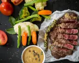 Un steak tranché, servi avec des légumes et deux types de sauces. L'une est une sauce crémeuse aux grains de poivre vert, l'autre est une sauce plus claire dans un bol.