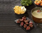 Un morceau de viande est trempé dans un caquelon de fondue. Des morceaux de viande, du brocoli, du chou-fleur et des légumes racines sont disposés sur une table en bois sombre.