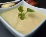 Un bol de beurre blanc crémeux, garni d'une feuille de persil. Des échalotes et un morceau de pain sont visibles en arrière-plan.